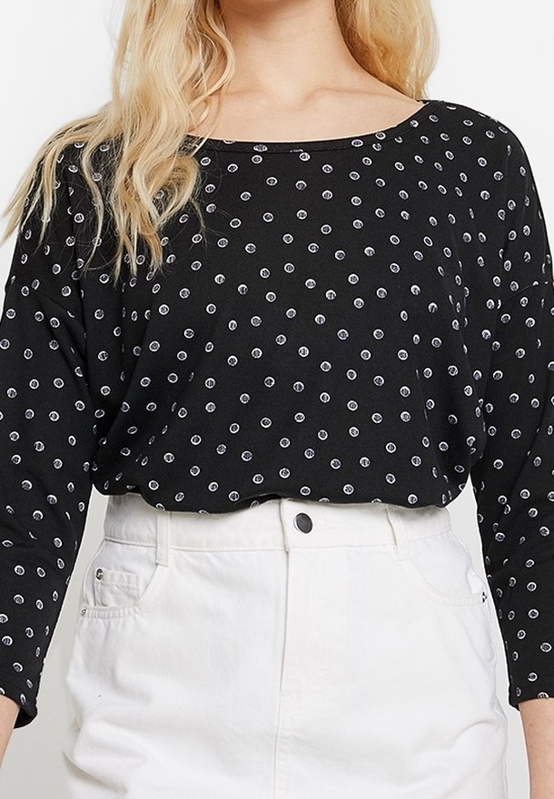 Schwarzes langärmliges Oberteil mit weißen runden Punkten, mit weitem Halsausschnitt und weichem Stoff. Kombiniert mit hochtaillierten weißen Shorts.