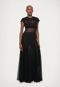 Črna čipkasta obleka s prosojnimi elementi, kapicami in raztegnjeno krilo. Ima teksturirane čipkaste podrobnosti in prozoren del bodice.