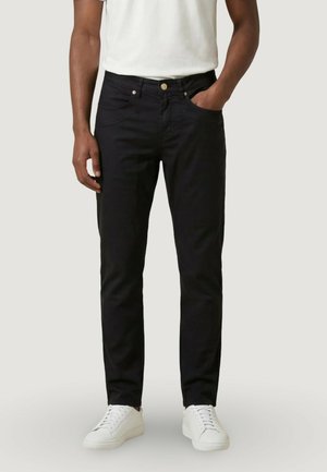 Uomo che indossa jeans neri slim fit, sneakers bianche e una t-shirt bianca, con una mano in tasca, in piedi davanti a uno sfondo neutro.