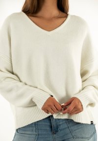Donna che indossa un dolcevita bianco a V e jeans blu, con le mani unite vicino all'orlo, mostrando anelli e una delicata collana.