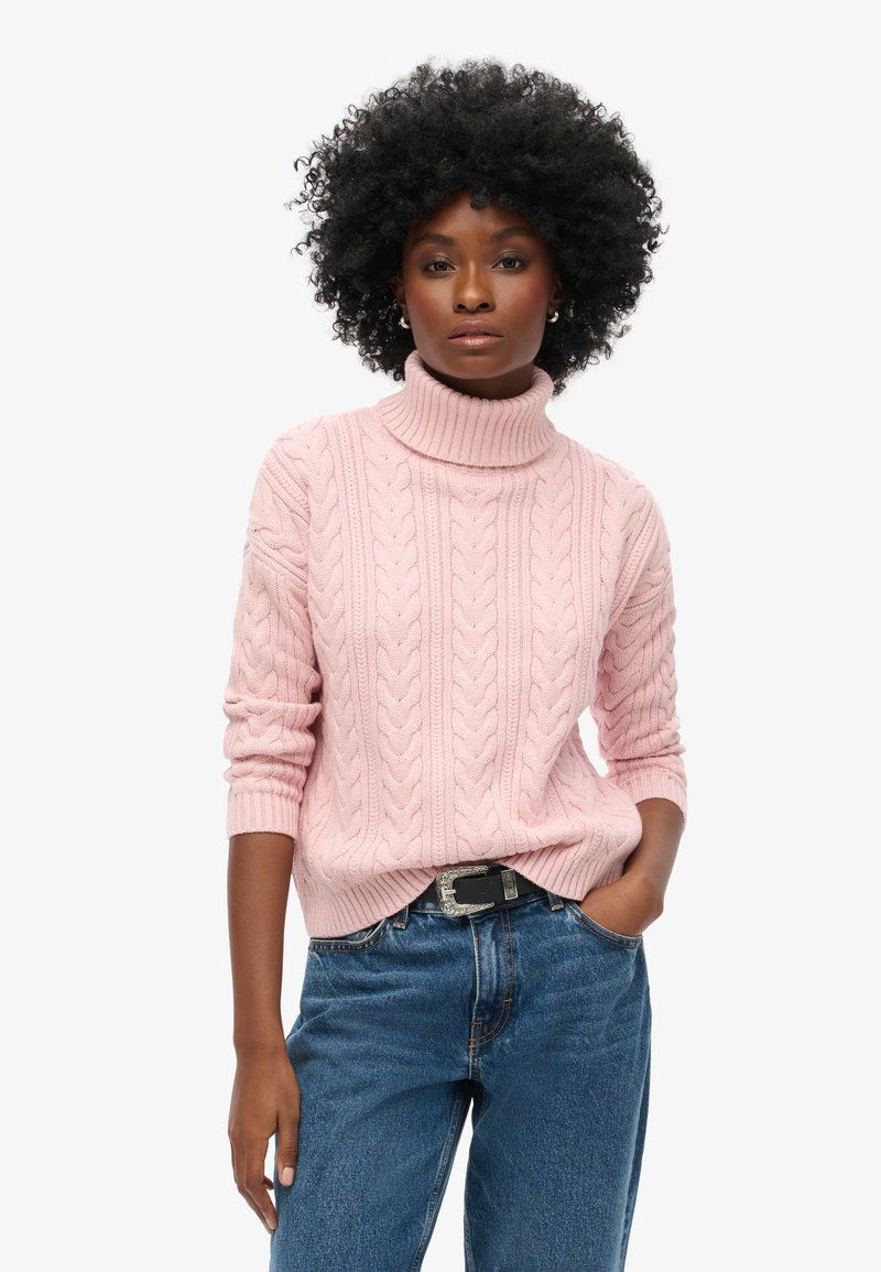 Lichtroze kabelgebreide turtleneck trui met driekwartmouwen, gecombineerd met blauwe denim jeans en een decoratieve zwarte riem.