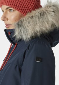 Navy winter jacket with fur-lined hood, red drawstrings, and black hardware accents. Features a logo patch on the sleeve.