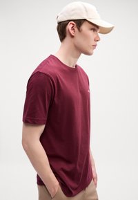 Jonge man in een beige pet en een kastanjebruine t-shirt met zijn handen in zijn zakken, staand met het gezicht naar rechts tegen een plain witte achtergrond.