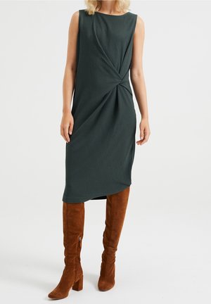 Mørkegrøn ribstrikket kjole til knæet med et snoet designdetalje ved taljen, kombineret med knæhøje brune ruskindsstøvler.