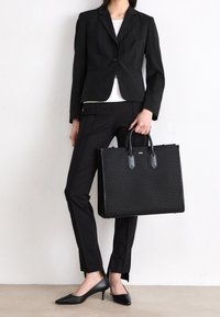 Black leather tote bag with textured surface and structured shape, paired with a black blazer, white top, and tailored black pants.