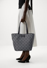 Borsa a tracolla bianca e nera con pois, dotata di manici robusti, realizzata in materiale sintetico. Presenta un'etichetta sul davanti.