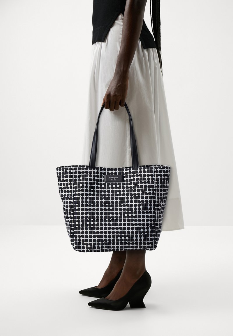 Borsa a tracolla bianca e nera con pois, dotata di manici robusti, realizzata in materiale sintetico. Presenta un'etichetta sul davanti.
