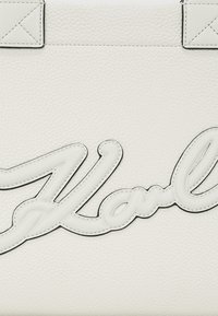 Balta texturēta ādas soma ar paceltu melnu izšūtu logo, kas raksta "Karl." Gludas akcenti uzlabo kopējo dizaina detaļu.