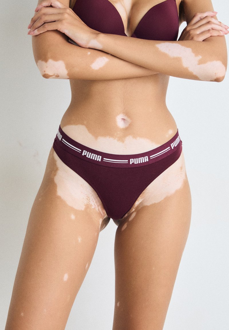 Vrouw met vitiligo in maroon Puma ondergoed en beha, met haar armen over haar borst gekruist voor een effen achtergrond.