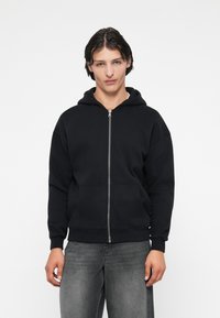 Melnā kapucēta džinsa jaka, izgatavota no kokvilnas sajaukuma, ar priekšējo rāvējslēdzēju, divām sānu kabatām un ribotām piedurknēm un apakšmalu. Modelis valkā pelēkos džinsus.