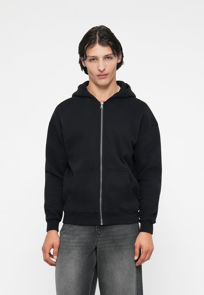 Melnā kapucēta džinsa jaka, izgatavota no kokvilnas sajaukuma, ar priekšējo rāvējslēdzēju, divām sānu kabatām un ribotām piedurknēm un apakšmalu. Modelis valkā pelēkos džinsus.