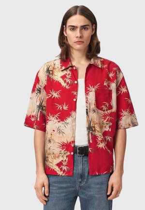 Młoda osoba o włosach do ramion, ubrana w czerwoną koszulkę z krótkimi rękawami z nadrukiem tygrysa i bambusa, biały top na ramiączkach, niebieskie dżinsy i czarny pasek.