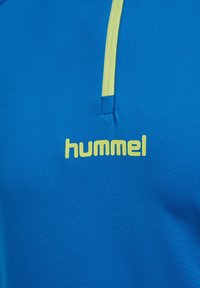 Blauwe sportshirt met contrasterend gele ritsdetail en "hummel" logo in het geel onderaan. Gladde textuur en aansluitend ontwerp.