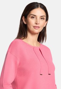 Różowy sweter z długim rękawem, okrągłym dekoltem i dwoma dekoracyjnymi sznurkami. Miękka, teksturowana tkanina o luźnym kroju.