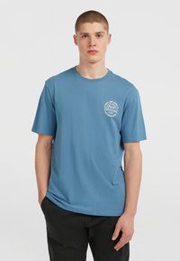 T-shirt en coton bleu clair à manches courtes, col rond, avec un graphique circulaire imprimé sur la zone du côté gauche de la poitrine. Pantalons noirs.