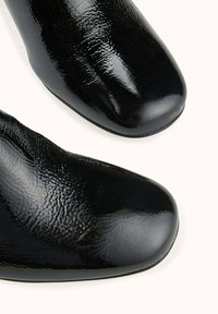 Gros plan sur les embouts en cuir noir brillant d'une paire de chaussures présentant une texture légèrement ridée sur un fond clair.