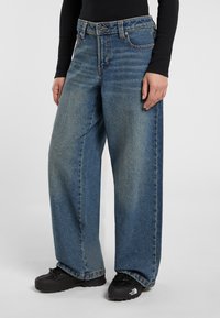 Person wearing loose-fitting blue denim jeans, black long-sleeve top, and black outdoor shoes with visible logo.