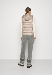 Chaleco acolchado sin mangas en beige claro, con capucha, acolchado horizontal y textura suave y brillante. Lleva puesto sobre un suéter crema.