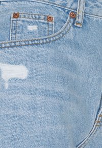 Tela vaquera azul claro con un diseño desgastado y bordes deshilachados. Incluye dos botones de tono cobre y un pequeño bolsillo parche.