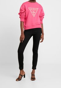 Różowa bluza z trójkątnym logo, zestawiona z czarnymi dopasowanymi dżinsami i czarnymi sandałami na obcasie. Gładkie tekstury i dopasowany krój.