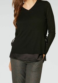 Pull noir à col en V avec manches longues et liens latéraux, superposé à un haut en satin sombre, assorti à un pantalon à carreaux texturé.
