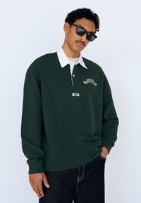 Grøn langærmet poloshirt med hvid krave, med frontlogo og et mærke. Brugt sammen med mørke jeans, vist mod en lys baggrund.