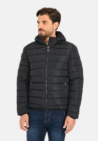 Uomo con capelli corti scuri e barba, indossa una giacca a vento nera trapuntata con cappuccio sopra dei jeans blu scuro, guarda verso sinistra.
