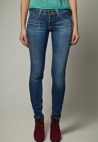 Úzké džíny v tmavě modré denimové barvě s vybledlým finišem, mají pět kapes a zapínání na knoflík. Nosené s burgundskými kotníkovými botami.