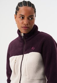 Fleecejacke mit lila Oberteil und cremefarbenem Unterteil. Hoher Kragen mit Frontreißverschluss, strukturierter Stoff und kleines Logodetail.