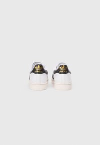 Bela Adidas čevlji z črnimi teksturiranimi poudarki na peti in zlatim logotipom. Nizka zasnova z gumijastim podplatom in šivanimi detajli.