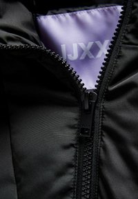 Tuva melnā džempera rāvējslēdzis daļēji atvērts, atklājot purpura audumu ar "JJXX" logo iekšpusē.