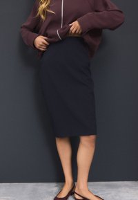 Jupe crayon bleu marine en tissu lisse ; présente une coupe ajustée, une longueur jusqu'au genou et des détails discrets sur la couture latérale. Portée avec un sweat-shirt sombre.