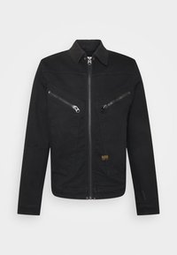 Zwarte lange mouwen jas met ritssluiting aan de voorkant, twee diagonale borstzakken met ritssluiting, kraag en een klein gouden merkje aan de voorkant.