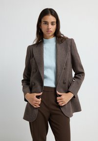 Blazer marrone a spina di pesce con design doppiopetto, abbinato a un dolcevita a costine blu chiaro e pantaloni tailleur marroni.