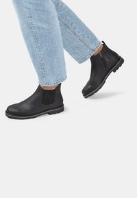 Stivali alla caviglia in pelle nera con pannelli laterali elasticizzati, punta arrotondata e suola in gomma, abbinati a jeans leggermente blu con risvolto.