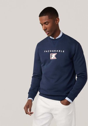 Marineblaue Sweatshirt mit gerippten Bündchen und Halsausschnitt, mit weißem Text und Logo auf der Brust, getragen über einem hellblauen kragenlosen Hemd.