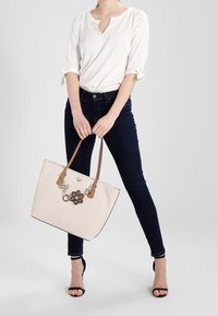Beżowa torba na ramię z brązowymi skórzanymi uchwytami i dekoracyjnymi detalami. Nosię ją z białą bluzką i ciemnymi dżinsami, na neutralnym tle.
