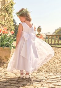 Ragazza giovane in un vestito bianco con un grande fiocco, che indossa una corona di fiori, tiene un cestino, in piedi su un sentiero di pietra vicino ai tulipani all'aperto.