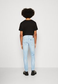 Person med lockigt hår som bär en svart T-shirt, ljusblå jeans och svarta stövlar, stående vänd mot en enfärgad vit vägg.