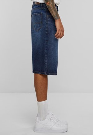 Gonna in denim a metà polpaccio di colore blu scuro con vestibilità regolare. Presenta una tasca posteriore ed è mostrata con sneakers bianche e calze a lunghezza media.