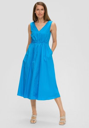 Blaues ärmelloses Kleid mit V-Ausschnitt, tailliertem Bund und Taschen. Glatte Textur und Midi-Länge. Modell kombiniert mit beigen Riemchensandalen.