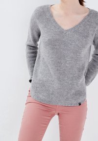 Kobieta w szarej, długoshowarowej bluzie typu V-neck i różowych spodniach, pozująca z rękami za plecami na białym tle.