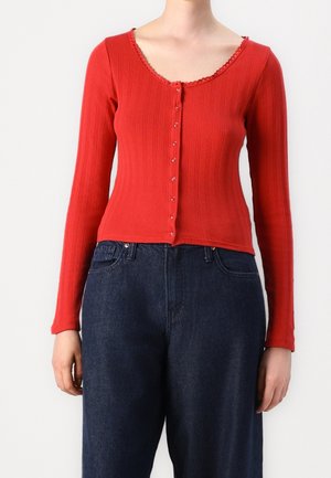Woman wearing a fitted, ribbed red buttoned cardigan with scalloped neckline and dark blue high-waisted jeans.