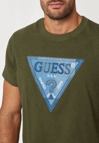 Alyvuogių žalios spalvos medvilnės marškinėliai su mėlyna trikampio grafika, kurioje pavaizduotas "GUESS" logotipas, tekstas ir klausimo ženklas, su tekstūra, primenančia džinsinį efektą.