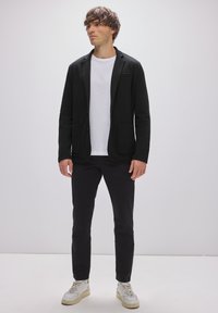 Blazer negro con dos bolsillos, llevado sobre una camiseta blanca y pantalones negros. El calzado incluye zapatillas blancas con detalles texturizados.