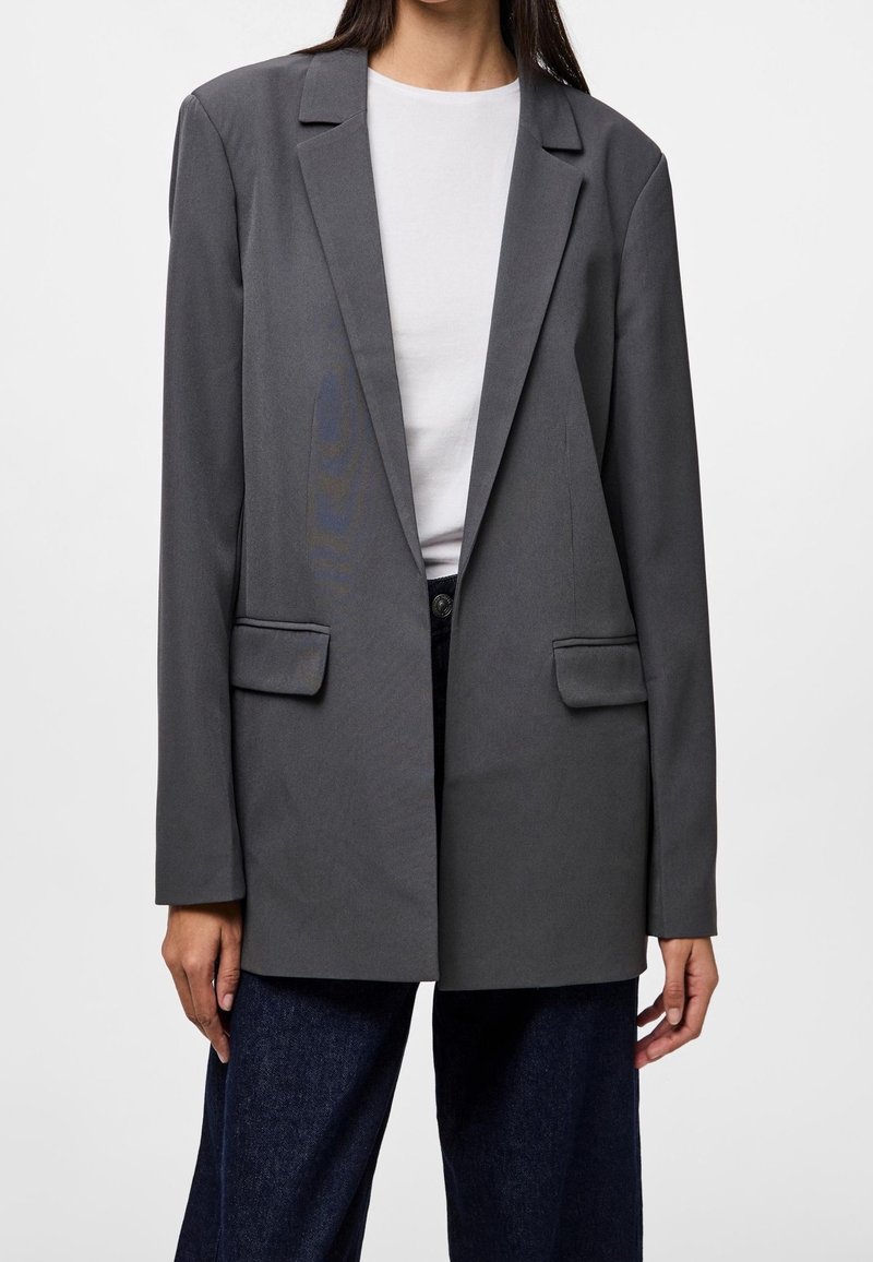 Blazer gris à coupe ajustée, avec revers crantés, poches poitrine et latérales, fabriqué en tissu lisse, associé à une chemise blanche simple.