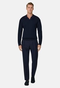 Homme portant un polo à manches longues bleu marine foncé, un pantalon assorti et des mocassins noirs, debout devant un fond uni.