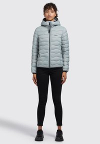 Lys blå pufferjakke med hætte. Har bølget syning, lynlås lukning og elastiske manchetter. Sammen med sorte bukser og sneakers.