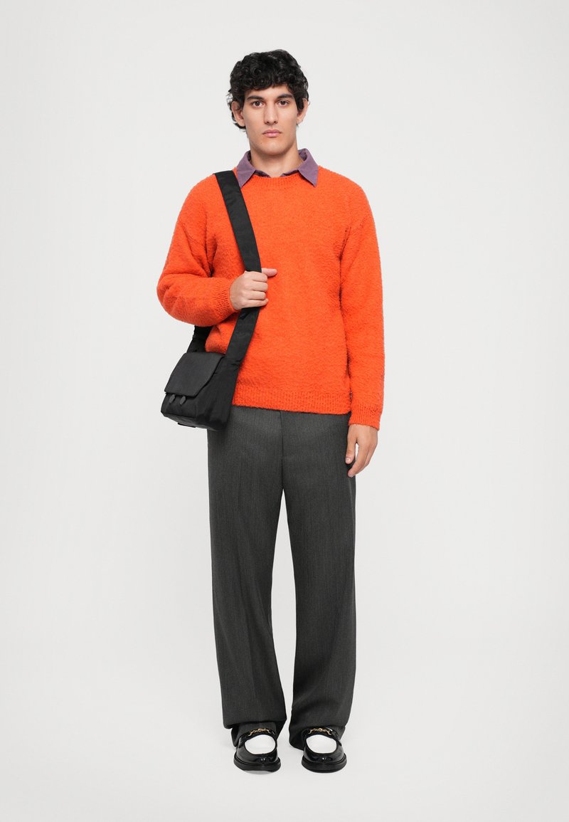 Oranges strukturiertes Sweatshirt, graue maßgeschneiderte Hose und schwarze Umhängetasche. Das Model trägt schwarz-weiße Schuhe und ist mit einem lila Kragenhemd geschichtet.