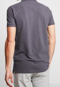 Polo shirt gris foncé à manches courtes, doté d'un col classique et d'un tissu lisse et texturé. Coupe ajustée avec des fentes sur les côtés à l'ourlet.